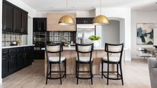 black kitchen with some light wood wall units and island, black and gray backsplash tiles, black bar stools, brass pendant lights, hardwood floor, white countertops, Living