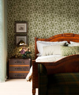 A bedroom with a green floral wallpaper, a nightstand with flowers on top of it, and a dark brown wooden bed with white sheets on top of it