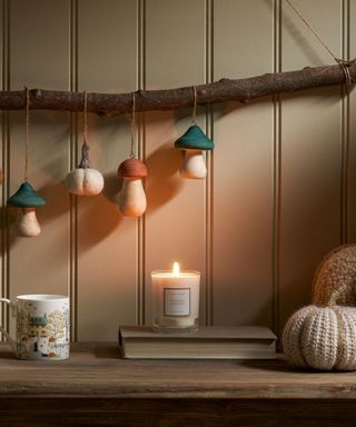 A kitchen corner with sage green paneling, a tree branch with mushroom decorations hanging from it, and a wooden countertop with a candle, mug, a book, and two fabric pumpkins