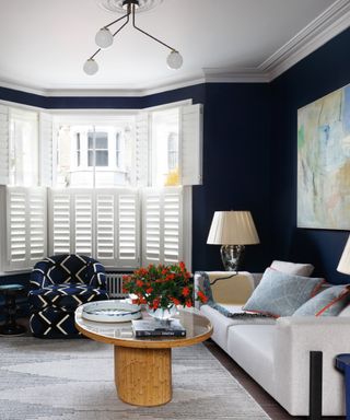 dark blue living room with white shutters, round coffee table, large painting, white couch, table lamps, retro pendant