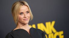 Reese Witherspoon with her blonde hair tied back wearing a black dress, with a black background with yellow writing behind her