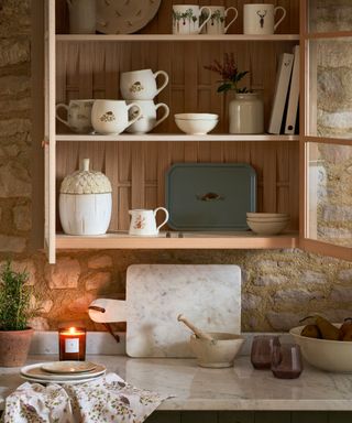 A kitchen pantry area with a wooden shelf unit with mugs inside of it and a kitchen counter with a marble board and a candle on top of it