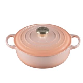 A round peach pink Le Creuset Dutch oven with handles on the side and a gold handle