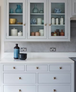 pale blue kitchen with pale blue cabinetry and glazed wall unit, white countertops, gray textured wallpaper backsplash,