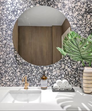 bathroom with terrazzo tiles, brass faucet, white countertop, round mirror, plant