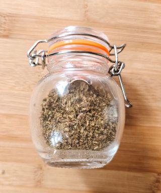 A small glass jar with dried basil inside of it, lying down on a wooden chopping board