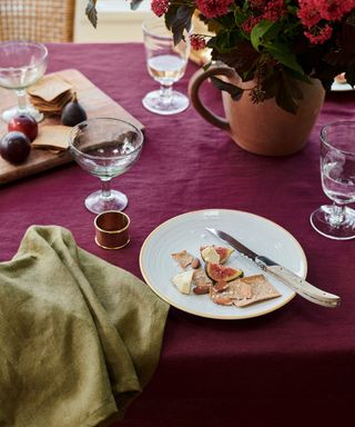 A purple dining table with a white plate, a vase of red flowers, an olive green cloth, and a cheeseboard on top of it