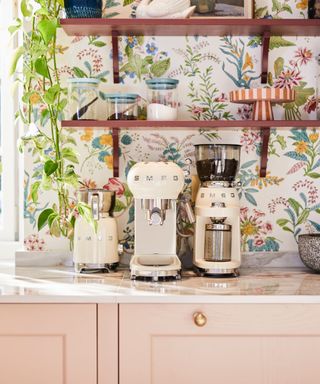 A marble kitchen countertop with three SMEG coffee makers, two wooden shelves with decor on, floral printed wallpaper, and light pink cabinets