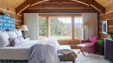 Farmhouse bedroom ideas are so chic. Here is one with a blue floral bed with white sheets and a throw pillow, rich wooden walls around it, a large window with long white curtains either side, and a burgundy armchair