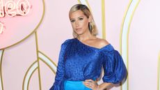 Ashley Tisdale wearing a blue dress with her hand on her hip, in front of a light pink, gold, and white background