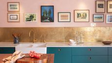 A kitchen with pink walls with wall art, a gold splashback, dark blue cabinets, and a wooden kitchen island with a chopping board with tomatoes on it