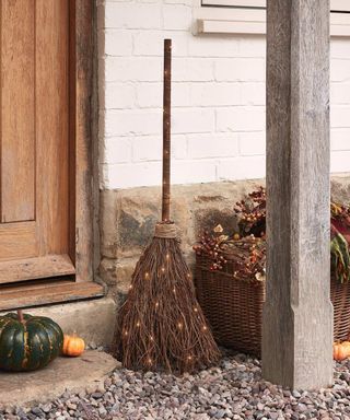 A front porch with a brown broomstick, pumpkins, and a basket on the stone gravel, with a wooden beam stood above it