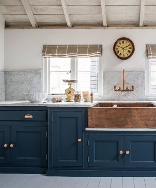 navy kitchen with blue veined marble countertops and backsplash, copper sink, blinds, white painted floor boards, copper faucet, clock, wooden ceiling,