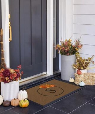 A fall front doorstep with a gray door and gray tiles with a doormat, pumpkins, and dried flowers on it