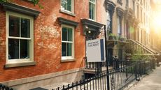 The outside of a traditional Brooklyn, New York, apartment building taken from the sidewalk at sunset. There is a white and blue sign hanging outside, which says 'Apartment for rent'