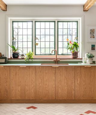 A kitchen with dark green grid windows, a green countertop with wooden cabinets underneath and a copper sink on top, and herringbone white flooring underneath